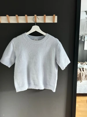 Ljusblå stickad t-shirt - Säljer en ljusblå stickad t-shirt med korta ärmar och rund halsringning. Tröjan har en avslappnad passform och ribbade kanter vid hals, ärmslut och nederkant. Perfekt för lager på lager och en stilren look.