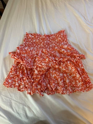 Orange blommig volangkjol - Supersöt kort kjol i orange med vitt blommönster och volangdetaljer. Kjolen har bred smock i midjan för skön passform och är gjord i ett lätt, luftigt material som passar perfekt till varma dagar.
