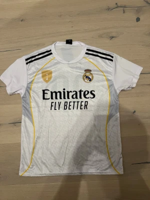 Real Madrid Mbappé matchtröja - Säljer en vit Real Madrid matchtröja med svarta och gula detaljer. Framsidan har klubbmärke, sponsorlogga och guldsköld. Baksidan har namnet 'Mbappé' och nummer 10 i svart. Tröjan har rund hals och korta ärmar, perfekt för fotbollsträning eller match.