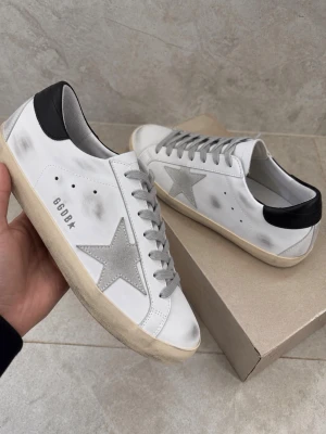 Golden Goose vita sneakers med stjärna - Golden Goose sneakers i vitt skinn med grå mockastjärna på sidan, svarta hälpartier och grå snörning. Klassisk låg modell med slitna detaljer för en cool vintage-look. Sulan är beige och skon har GGDB-tryck på sidan.
