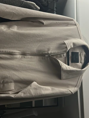Beige tunn jacka från jack and Jones - Snygg beige jacka med minimalistisk design och dragkedja framtill. Jackan har ståkrage, långa ärmar och två fickor framtill. Tillverkad i ett mjukt, tunt material som passar perfekt för lager-på-lager. Enkel och stilren look. Ingen Bruksmärka