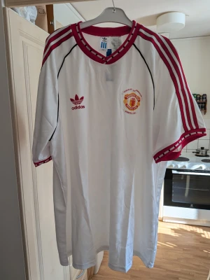 Vit Manchester United Adids retrotröja - Säljer en vit Manchester United retrotröja från Adidas med röda detaljer och klubbmärke på bröstet. Tröjan har röda ränder på axlarna, svart piping och rund halsringning med röd kant. Perfekt för fotbollsfans som gillar klassisk stil. Storlek XXXL