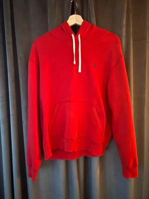 Röd Ralph Lauren hoodie - Snygg röd hoodie med huva och vita dragsnören. Klassisk magficka framtill och ribbade muddar vid ärmar och nederkant. Liten broderad logga på bröstet. Perfekt för en chill och avslappnad stil.