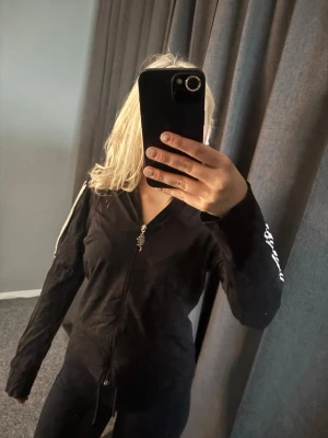 Svart hoodie från Urban Wear - Säljer en svart hoodie från Urban Wear med dragkedja framtill och coolt metallhänge på zippen. Tröjan har huva och vita detaljer på ärmarna samt tryck på ena ärmen. Perfekt för en avslappnad streetstyle-look. Liten i storleken!