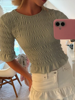 Ljusgrön smockad topp med volang - Superfin ljusgrön topp med smockad struktur och volangkant nertill. Toppen har rund halsringning och halvlånga ärmar. Perfekt att styla med kjol eller jeans för en fräsch look. Skulle säga att färgen är åt det pastell gröna hållet, superfint och somrigt!!