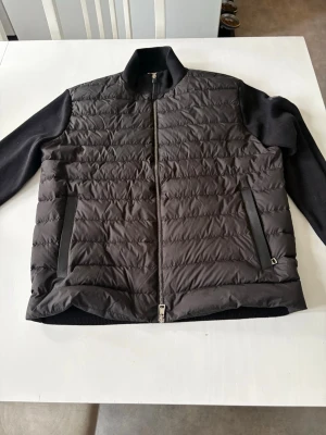 Moncler cardigan black label - Snygg svart pufferjacka med quiltad front och rygg samt stickade ärmar. Jackan har hög krage, dragkedja framtill och två fickor med dragkedja. Perfekt för kalla dagar när du vill hålla stilen och värmen.