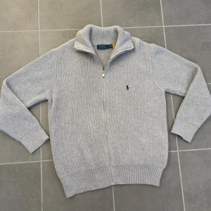 Grå ribbad kofta från Polo Ralph Lauren - Säljer en grå ribbstickad kofta från Polo Ralph Lauren med halv dragkedja och hög krage. Klassisk marinblå logga broderad på bröstet. Perfekt till höst och vinter, riktigt skön och stilren. Passar dig som gillar en clean look.