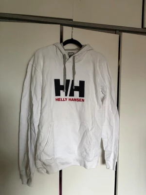 Vit hoodie från Helly Hansen - Säljer en vit hoodie från Helly Hansen med stor svart HH-logga och röd text på bröstet. Hoodien är inne i huvan gråfärgad