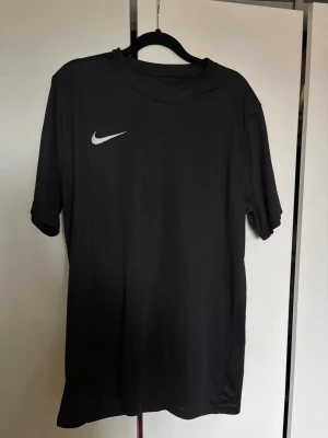 Svart Nike Dri-Fit träningströja  - Svart sportig träningströja från Nike. Tröjan har korta ärmar, rund halsringning och en vit Nike-logga på bröstet. Perfekt för gymmet eller löprundan.