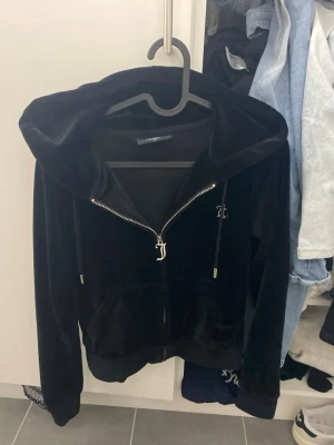 Svart velour hoodie från Juicy Couture - Säljer en svart velour hoodie från Juicy Couture med dragkedja framtill och fickor. Stor huva och snören med metalländar. På ryggen står det 'Juicy' i glittriga stenar. Perfekt för en chill och trendig look.