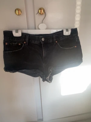 Svarta jeansshorts med rå kant - Säljer ett par svarta jeansshorts med rå kant nertill och klassisk femficksdesign. Shortsen har knapp och dragkedja framtill samt bälteshällor. Perfekta för varma dagar och ger en avslappnad look. (Liten i storlek)