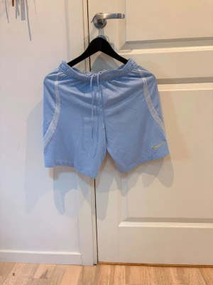 Ljusblå Nike shorts med snörning - Säljer ett par ljusblå shorts från Nike med vit detalj längs sidorna och snörning i midjan. Shortsen har en broderad Nike-logga i gult på vänster ben och är tillverkade i ett lätt och bekvämt material, perfekta för träning eller chill.