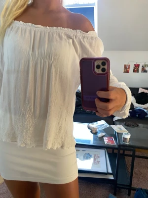 Vit offshoulder blus med spetsdetaljer - Säljer en vit offshoulder blus med långa ärmar och fina spetsdetaljer längst ner. Blusen har en luftig och flowy passform i ett tunt, skönt tyg som passar perfekt till sommaren. Elastisk kant upptill för en snygg passform över axlarna.