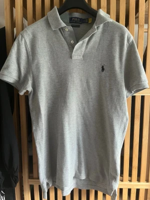 Grå pikétröja från Polo Ralph Lauren - Klassisk grå pikétröja från Polo Ralph Lauren med korta ärmar och krage. Tröjan har knappar vid halsen och den ikoniska broderade loggan på bröstet. Tillverkad i mjuk bomull som känns skön mot huden.