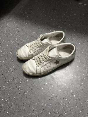 YSL (Yves Saint Laurent) - Säljer ett par vita sneakers med snörning och stjärndetaljer på sidorna. Skorna har en klassisk låg profil och är tillverkade i tyg eller syntetmaterial. Perfekta för dig som gillar en lekfull och avslappnad stil.