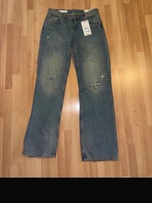 Blå raka jeans med slitningar - Säljer ett par blå jeans med raka ben och tydliga slitningar framtill och baktill. Jeansen har klassisk femficksmodell och är gjorda i denim. Perfekta för en avslappnad och trendig look.