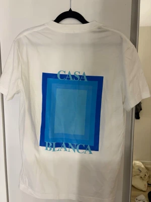 Vit t-shirt med blått tryck från Casablanca - Vit t-shirt från Casablanca med ett stort grafiskt tryck i olika blå nyanser och texten 'CASA BLANCA' på ryggen. Mindre liknande tryck på bröstet. Klassisk passform och rund hals. Perfekt för en clean och modern stil.