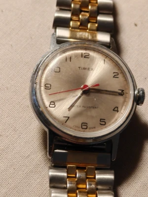 Vintage Timex Automatic - Stilren analog klocka från Timex med rund urtavla i silver och tydliga svarta siffror. Armbandet är i metall med både silver- och guldfärgade länkar. Klockan har tre visare, inklusive en röd sekundvisare, och en klassisk design som passar till många stilar.