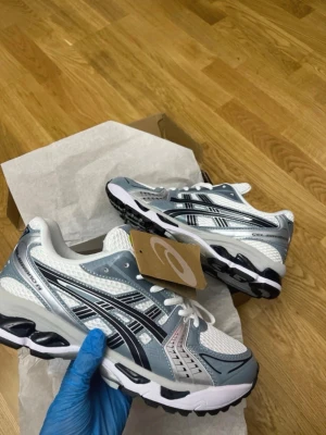 Asics Gel-Kayano 14 vita/grå sneakers - Snygga ljus blåa Asics Gel-Kayano 14 sneakers med vitt grått och svarta detaljer. Ovandelen är i mesh och syntet för bra andningsförmåga och komfort. Klassisk sportig siluett med vita snören och robust sula. Perfekta för dig som gillar retrostil och streetwear.