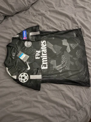 Svart PSG matchtröja Neymar Jr - Snygg svart PSG matchtröja med Neymar Jr och nummer 10 på ryggen. Tröjan har vita detaljer, tryckta loggor och mönster, samt Fly Emirates sponsortryck. UEFA-märke på ärmen och Paris Saint-Germain klubbmärke på bröstet.