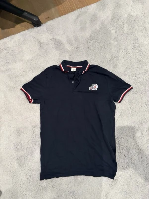 Marinblå pikétröja från Moncler - Snygg marinblå pikétröja från Moncler med klassisk krage och knappar framtill. Tröjan har vita och röda detaljer på krage och ärmslut samt Moncler-logga på bröstet. Tillverkad i mjuk bomull för en bekväm känsla.