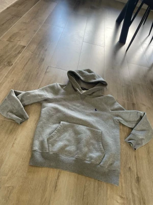 Grå polo Ralph lauren hoddie - Säljer en enkel och stilren grå polo Ralph lauren hoddie med stor känguruficka framtill och huva. Hoodien har en liten broderad logga på bröstet och är gjord i mjukt sweatshirtmaterial. Perfekt för en avslappnad och chill look.