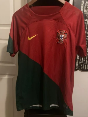 Portugal Nike fotbollströja röd/grön - Snygg Portugal fotbollströja från Nike i rött och grönt med gul Nike-logga och Portugals landslagsemblem på bröstet. Tröjan har korta ärmar och sportig passform, perfekt för match eller träning.