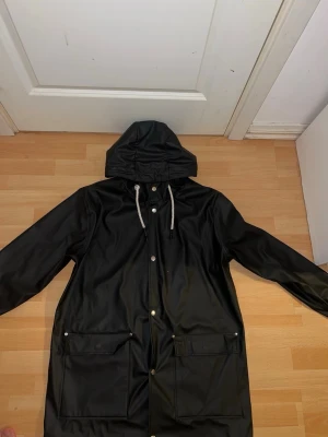 Svart regnjacka med huva - Snygg svart regnjacka med huva och vita dragsnören. Jackan har tryckknappar framtill och två stora fickor med lock. Tillverkad i vattenavvisande material med lång passform som skyddar mot regn. Perfekt för blöta dagar.