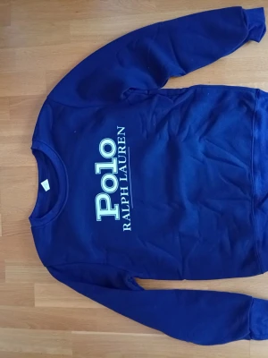 Mörkblå sweatshirt Polo Ralph Lauren - Mörkblå sweatshirt från Polo Ralph Lauren med stort vitt tryck på bröstet. Klassisk rund halsringning och långa ärmar. Perfekt för en avslappnad och stilren look.