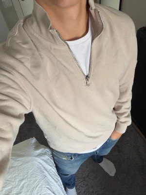 Beige halfzip tröja - Säljer en beige tröja med halv dragkedja och hög krage. Tröjan har en avslappnad passform och ribbade muddar vid ärmar och nederkant. Perfekt för en chill och stilren look. Materialet känns mjukt och bekvämt, troligen bomullsblandning.