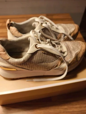 Glittriga sneakers i guld och vitt -  sneakers från Michael Kors i vitt och guld med glittriga detaljer. Skorna har vita snören, meshpartier och guldfärgade metalldetaljer. Slitna, har sina skavanker men är fullt fungerande 