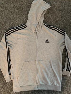 Grå zip hoodie från Adidas - Snygg grå hoodie från Adidas med dragkedja framtill och klassiska svarta ränder längs ärmarna. Huva med dragsko och kängurufickor. Adidas-logga broderad på bröstet. Tillverkad i mjukt sweatshirtmaterial, perfekt för chill eller träning.