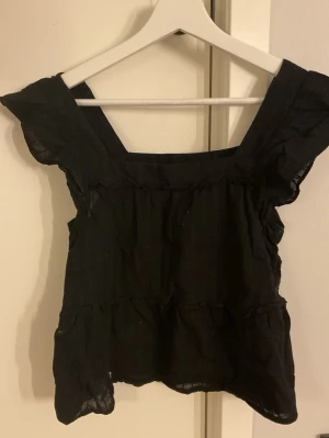 Svart topp - Snygg svart topp med fyrkantig ringning och volangärmar. Tillverkad i ett tunt, lätt tyg som ger en luftig känsla. Perfekt att matcha med jeans eller kjol för en trendig look.