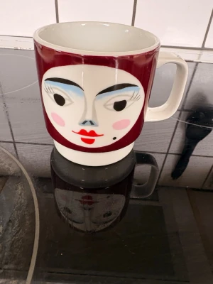 Mugg med ansiktsmotiv från B&G - Unik porslinsmugg från Bing & Grøndahl med ett färgstarkt ansiktsmotiv i rött, blått och svart. Muggen har vit bas och ett stort handtag, perfekt för dig som gillar retro eller dansk design. Märkt undertill med B&G Copenhagen Porcelain.