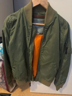 Olivgrön Alpha jacka med orange foder.  - Snygg olivgrön bomberjacka med klassiskt orange foder. Jackan har ribbade muddar vid krage, ärmslut och nederkant samt fickor med tryckknappar och en ärmficka med dragkedja. Tillverkad i ett glansigt syntetmaterial som ger en cool look.