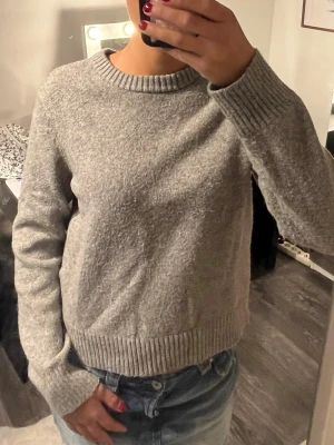 Grå stickad tröja HM - Mysig grå stickad tröja med rund halsringning och ribbade muddar vid ärmslut och nederkant. Tröjan har en enkel, rak passform och är perfekt att matcha med jeans för en avslappnad look. Materialet ser ut att vara mjukt och värmande.