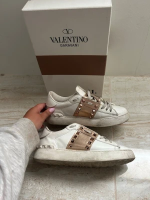 Valentino Garavani sneakers med nitar - EU 36, men passar mig som har storlek 37,5. Kommer med extra valentino slosnören, dustbagen & kartongen. Äkta vita sneakers från Valentino Garavani med breda metallicrosa band och guldiga nitar över vristen. Kan gå ner i pris vid snabbt köp. Köptes av en tjej på plick för två år sen. Skorna har en chunky sula och coola detaljer i läder och textil. 