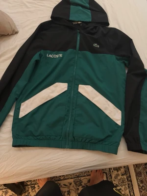 Grön och svart vindjacka från Lacoste - Snygg vindjacka från Lacoste i grön och svart färg med vita detaljer på fickorna. Jackan har huva, dragkedja och logotyp på bröstet. Perfekt för att hålla vinden borta och ge en sportig look. Priset kan diskuteras vid snabbaffär!