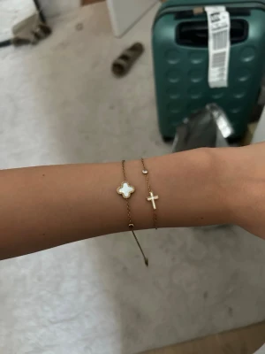 Guldfärgade armband med fyrklöver och kors - Säljer två tunna guldfärgade armband med en fyrklöverformad detalj i vitt, och ett kors. Kedjan är finlänkad och har en liten rund dekoration. Perfekt för dig som gillar stilrena och eleganta smycken.