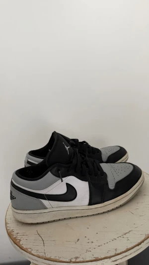 Nike Air Jordan 1 Low svart/grå - Snygga Nike Air Jordan 1 Low sneakers i svart, grått och vitt. Skorna har klassisk låg siluett, svart Swoosh och Jordan Wings-logga på hälen. Ovandelen är i läder med perforerad tåbox för extra ventilation. Perfekta för dig som gillar streetwear.