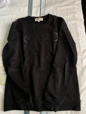 Svart Kenzo Paris sweatshirt - Säljer en svart sweatshirt från Kenzo med stor, upphöjd logga 'KENZO PARIS' på bröstet. Tröjan har rund halsringning, långa ärmar och ribbade muddar vid ärmslut och nederkant. Perfekt för dig som gillar stilrena och exklusiva plagg.
