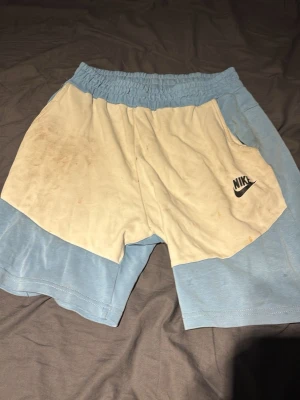 Nike blå och vita shorts - Säljer ett par blå och vita shorts från Nike med elastisk midja och sidoficka med dragkedja bak. Shortsen har en avslappnad passform och är tillverkade i mjukt material, perfekta för träning eller chill. Nike-logga på framsidan. Den har smuts på fast orkar inte tvätta bort de. Man kan bara lägga den i tvättmaskinen och tvätta de så går de bort 