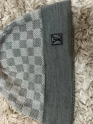 Gråmelerad mössa med rutmönster - Snygg gråmelerad mössa med diskret rutmönster och bredare enfärgad kant. Mössan har en liten broderad patch framtill. Tillverkad i mjukt stickat material som känns skönt mot huden. Perfekt för dig som vill ha en stilren och trendig accessoar.