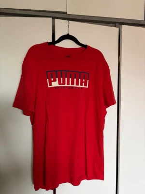 Röd PUMA t-shirt med tryck - Snygg röd t-shirt från PUMA med stort logotryck på bröstet i blått och vitt. Klassisk passform med korta ärmar och rund halsringning. 