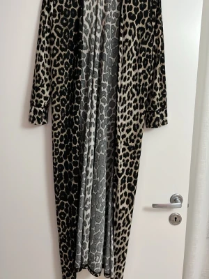 Lång leopardmönstrad morgonrock - Snygg och trendig lång morgonrock med leopardmönster i beige, svart och brunt. Morgonrocken har långa ärmar och öppen front, perfekt för att chilla hemma i stil. Materialet ser mjukt och bekvämt ut, vilket gör den extra skön att bära.
