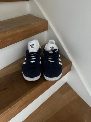 Adidas Gazelle mörkblå sneakers - Säljer ett par mörkblå Adidas Gazelle sneakers i mocka med klassiska vita ränder och vit sula. Använt skorna 2 gånger och de var för små därför så säljs de. 