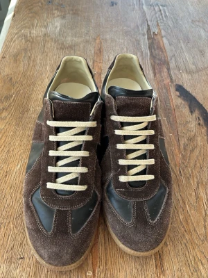 Bruna sneakers i mocka och läder Masion Margiela storlek 42 - Snygga sneakers i mörkbrun mocka och svart läder med beige snörning och gummisula. Klassisk låg modell med retrovibbar och neutrala färger som passar till det mesta. Perfekt för dig som gillar stilrena och tidlösa skor. Inköpta för runt 6000 