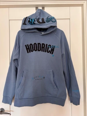 Blå hoodie från Hoodrich med tryck strl L  - Säljer en blå hoodie från Hoodrich med svart och blått tryck på bröstet och huvan. Hoodien har stor magficka, huva med broderad text och tryck även på ryggen. Tillverkad i mjukt bomullsmaterial och har ribbade muddar vid ärmar och nederkant.