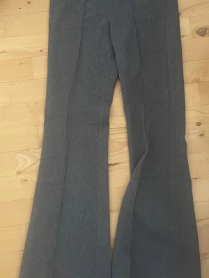 Gråa utsvängda kostymbyxor - Snygga grå kostymbyxor med utsvängda ben och pressveck framtill. Byxorna har en klassisk midja och är tillverkade i ett mjukt, vävt material som ger en stilren look. Perfekta för dig som vill ha en trendig och bekväm byxa.