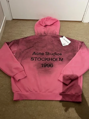 Rosa hoodie Acne Studios Stockholm - Säljer en rosa hoodie från Acne Studios med svart text 'Acne Studios STOCKHOLM 1996' på ryggen. Hoodien har en unik tvättad look med mörkare partier och klassisk huva. Perfekt för dig som gillar streetwear och vill sticka ut.